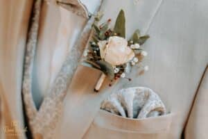 Nahaufnahme einer hellbeigen Anzugsjacke mit einer weißen Rosenboutonniere und Grünzeug am Revers. Ein gemustertes Einstecktuch ist ordentlich in die Brusttasche gesteckt.