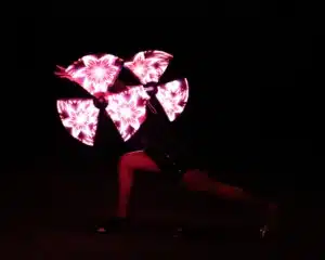 Ein Darsteller in Ausfallschritt-Pose dreht vier LED-Fächer, die helle, symmetrische rosa Lichtmuster vor einem dunklen Hintergrund erzeugen.