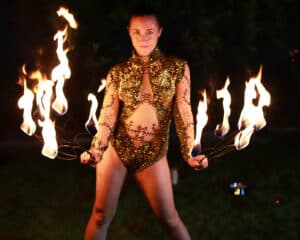Ein Darsteller in einem glitzernden goldenen Kostüm hält mehrere brennende Fackeln in jeder Hand, die Feuerbögen in der Nacht erzeugen, mit einem Hintergrund aus verschwommenem Grün.