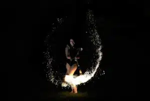 Ein dunkel gekleideter Darsteller wirbelt nachts Feuerpoi und erzeugt einen hellen, funkelnden Lichtbogen vor einem dunklen Hintergrund.