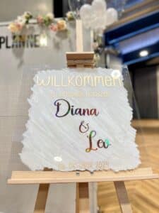 Auf einem dekorativen Willkommensschild auf einer Staffelei steht "Willkommen zu unserer Hochzeit Diana & Leo 06. Oktober 2024" in deutscher Sprache vor einem unscharfen Hintergrund mit Blumen- und Ballondekorationen.