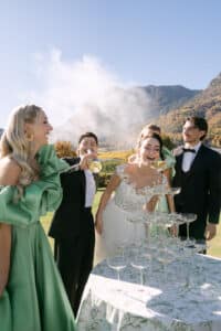 Braut und Bräutigam lachen mit Freunden im Freien bei einem Sektturm, während ein Gast Champagner einschenkt. Die Gruppe ist förmlich gekleidet, mit Bergen und Herbstbäumen im Hintergrund unter einem klaren Himmel.