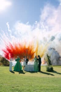 Eine Gruppe von formell gekleideten Personen steht im Gras in der Nähe eines Tisches, während im Hintergrund unter einem klaren blauen Himmel bunte Feuerwerkskörper und Rauch explodieren.