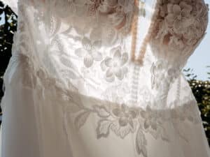 Nahaufnahme eines Hochzeitskleides aus weißer Spitze mit Blumenmustern, beleuchtet vom Sonnenlicht. Die filigranen Details der Spitze sind deutlich sichtbar, und das Kleid scheint im Freien zu hängen, mit etwas Grün im Hintergrund.