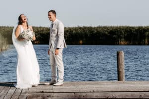 Eine Braut in einem weißen Kleid und Schleier lacht, während sie Blumen in der Hand hält. Sie steht neben einem Bräutigam in einem hellen Anzug auf einem hölzernen Steg neben einem See mit hohem Schilf im Hintergrund.