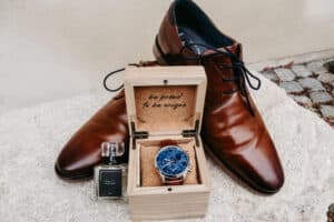 Ein Paar braune Lederschuhe, ein Holzkästchen mit einer blauen Armbanduhr und der Aufschrift "be proud to be unique" sowie ein Fläschchen Kölnisch Wasser stehen auf einer hellen Steinfläche.