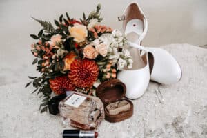 Ein flacher Brautschuh, ein bunter Blumenstrauß, ein Parfümfläschchen, ein Lippenstift, ein offenes hölzernes Ringkästchen mit zwei Ringen und verstreute Perlen sind auf einer hellen Steinoberfläche angeordnet.