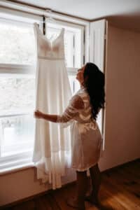Eine Frau in einem weißen Gewand steht an einem Fenster und hält ein weißes, ärmelloses Hochzeitskleid hoch, das am Fensterrahmen hängt. Sanftes Tageslicht strömt durch das Fenster und beleuchtet die Szene.
