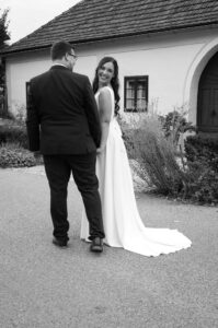 Eine lächelnde Braut in einem langen weißen Kleid hält die Hand ihres Partners in einem dunklen Anzug, während sie vor einem Haus spazieren gehen und in die Kamera schauen. Die Szene ist in schwarz-weiß gehalten.