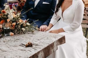 Eine Braut in einem weißen Kleid und ein Bräutigam in einem marineblauen Anzug sitzen Seite an Seite und halten sich an einem rustikalen Holztisch, der mit einem Blumenstrauß geschmückt ist. Nur ihre Torsos und Hände sind zu sehen.
