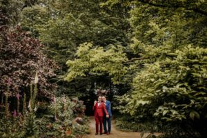 Zwei Männer in Anzügen, ein roter und ein blauer, stehen dicht beieinander auf einem Gartenweg, umgeben von üppig grünen Bäumen und blühenden Pflanzen, lächeln und halten sich an den Händen.