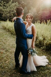 Eine Braut in einem weißen Spitzenkleid mit einem Blumenstrauß in der Hand steht mit ihrem Bräutigam in einem marineblauen Anzug im Freien, umgeben von Grünpflanzen und sanftem Sonnenlicht, das durch die Bäume fällt.