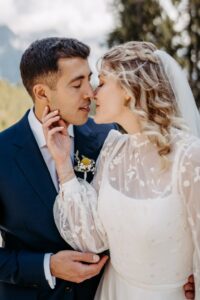 Eine Braut und ein Bräutigam stehen im Freien dicht beieinander und wollen sich küssen. Die Braut trägt ein weißes Kleid mit durchsichtigen Blumenärmeln und einen Schleier, während der Bräutigam einen dunklen Anzug mit einer Ansteckblume trägt. Im Hintergrund sind Bäume und Berge zu sehen.