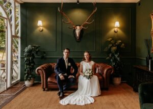 Eine Braut in einem weißen Kleid und ein Bräutigam in einem dunklen Anzug sitzen auf einem braunen Ledersofa in einem grünen Raum, der mit Pflanzen, Wandleuchten und einem darüber angebrachten Hirschschädel mit Geweih dekoriert ist.