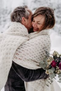 Ein in eine kuschelige weiße Decke gehülltes Paar umarmt sich im Freien im Schnee. Die Frau lächelt und hält einen Strauß lila Rosen und Grünzeug in der Hand, während Schneeflocken ihr Haar bestäuben.