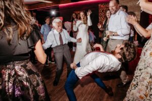 Eine lebhafte Szene eines Hochzeitsempfangs, bei dem die Gäste ausgelassen tanzen. Ein älterer Mann und ein jüngerer Mann im Anzug stehen in der Mitte, der jüngere Mann lehnt sich dramatisch zurück, während andere um sie herum klatschen und jubeln.