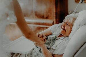 Eine ältere Frau liegt in einem Krankenhausbett und lächelt warm, während sie einem Besucher, der neben ihr steht, die Hand hält, was ein Gefühl von Trost und Verbundenheit vermittelt.