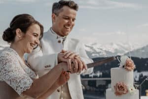 Ein lächelndes Paar in weißer Hochzeitskleidung schneidet eine mit rosa und weißen Blumen geschmückte Stufentorte an, während im Hintergrund verschneite Berge und ein See zu sehen sind.