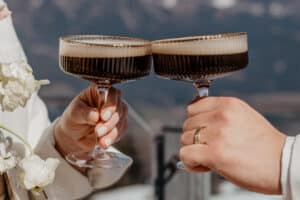 Zwei Hände, eine davon mit einem goldenen Ring, stoßen mit Vintage-Cocktailgläsern an, die mit einem dunklen, schaumigen Getränk gefüllt sind. Auf der linken Seite sind zarte weiße Blumen zu sehen, und im Hintergrund sind verschwommene Berge zu erkennen.