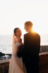 Ein Brautpaar steht eng beieinander, lächelt und hält Händchen, während hinter ihnen die Sonne über dem Meer untergeht. Die Braut trägt ein schulterfreies Kleid und einen Schleier, der Bräutigam einen dunklen Anzug.