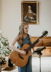 Eine Frau in einem blauen Kleid spielt Akustikgitarre und singt. Sie steht neben einer Pflanze und vor einer Holzbank. An der Wand hinter ihr hängt ein Gemälde einer historischen Figur.