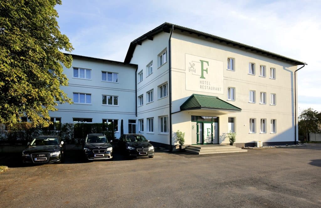 Ein dreistöckiges weißes Gebäude mit der Aufschrift „F Hotel Restaurant“ an der Fassade. Davor parken vier schwarze Autos. Der Eingang ist mit einem grünen Vordach überdacht und auf der linken Seite des Gebäudes steht ein großer Baum.